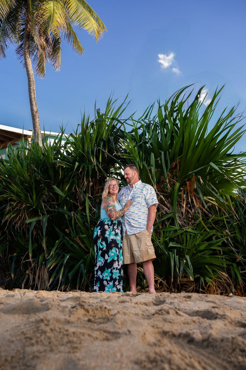 Couple embracing on sandy beach surrounded by tropical palm trees and lush green plants under a bright blue sky.
