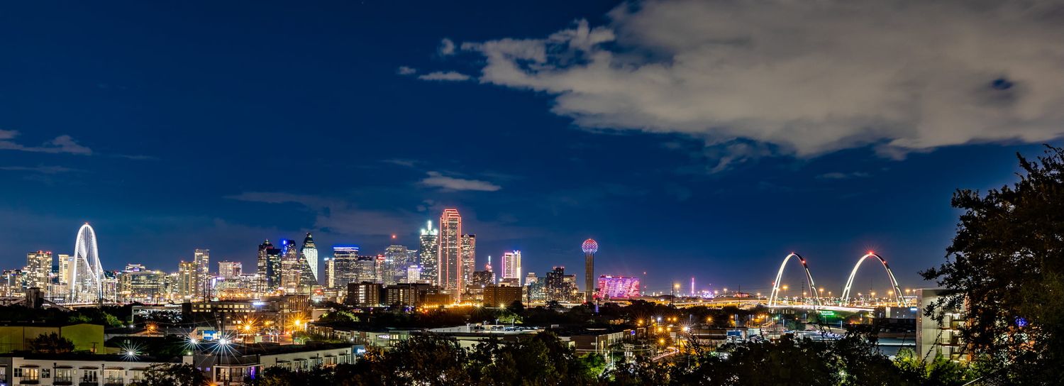 Panoramic nighttime skyline of Dallas, Texas, with illuminated Calatrava Gateway Arch and downtown buildings under dark blue sky.