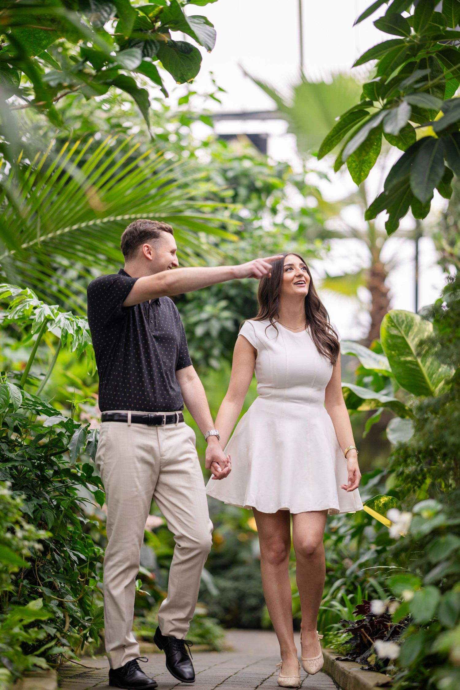 A couple walks together in a lush greenhouse garden setting with tropical plants and greenery surrounding them.