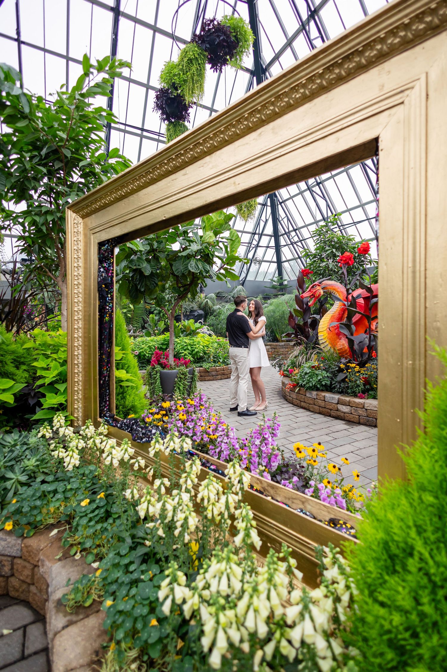 A large ornate gold frame creates a romantic scene in a conservatory filled with colorful flowers.