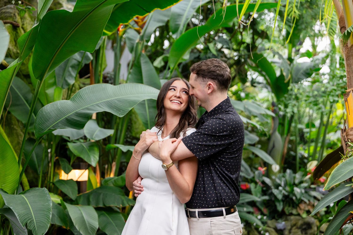 A romantic couple shares a tender moment in a lush tropical garden setting with vibrant green foliage.