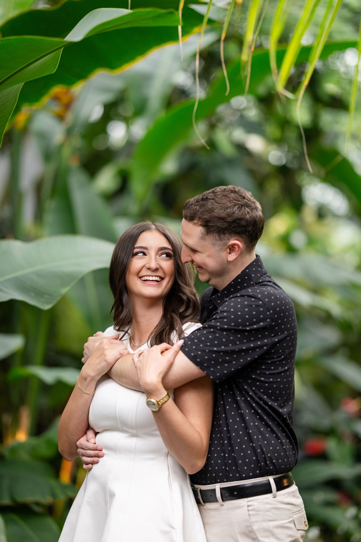 A couple shares a romantic moment together in a lush greenhouse setting with tropical greenery surrounding them.