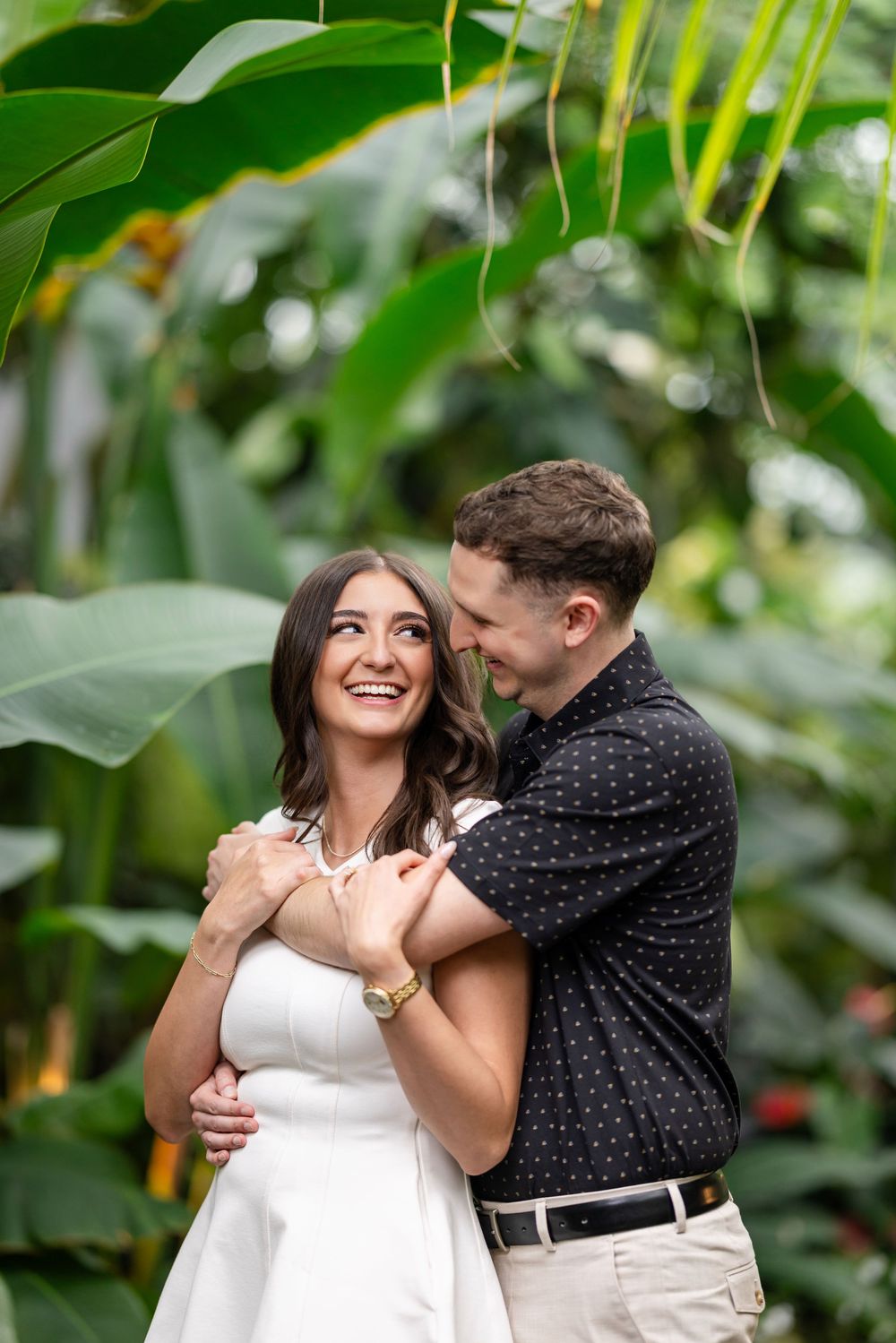 A couple shares a romantic moment together in a lush greenhouse setting with tropical greenery surrounding them.