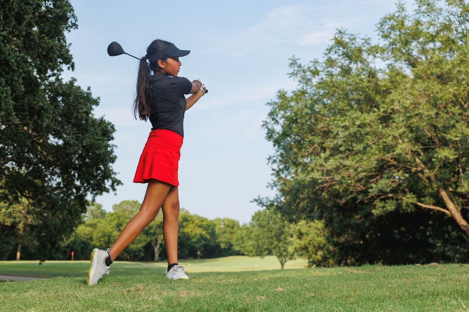 Young golfer in navy shirt and red shorts performs full swing on lush green golf course during summer day.