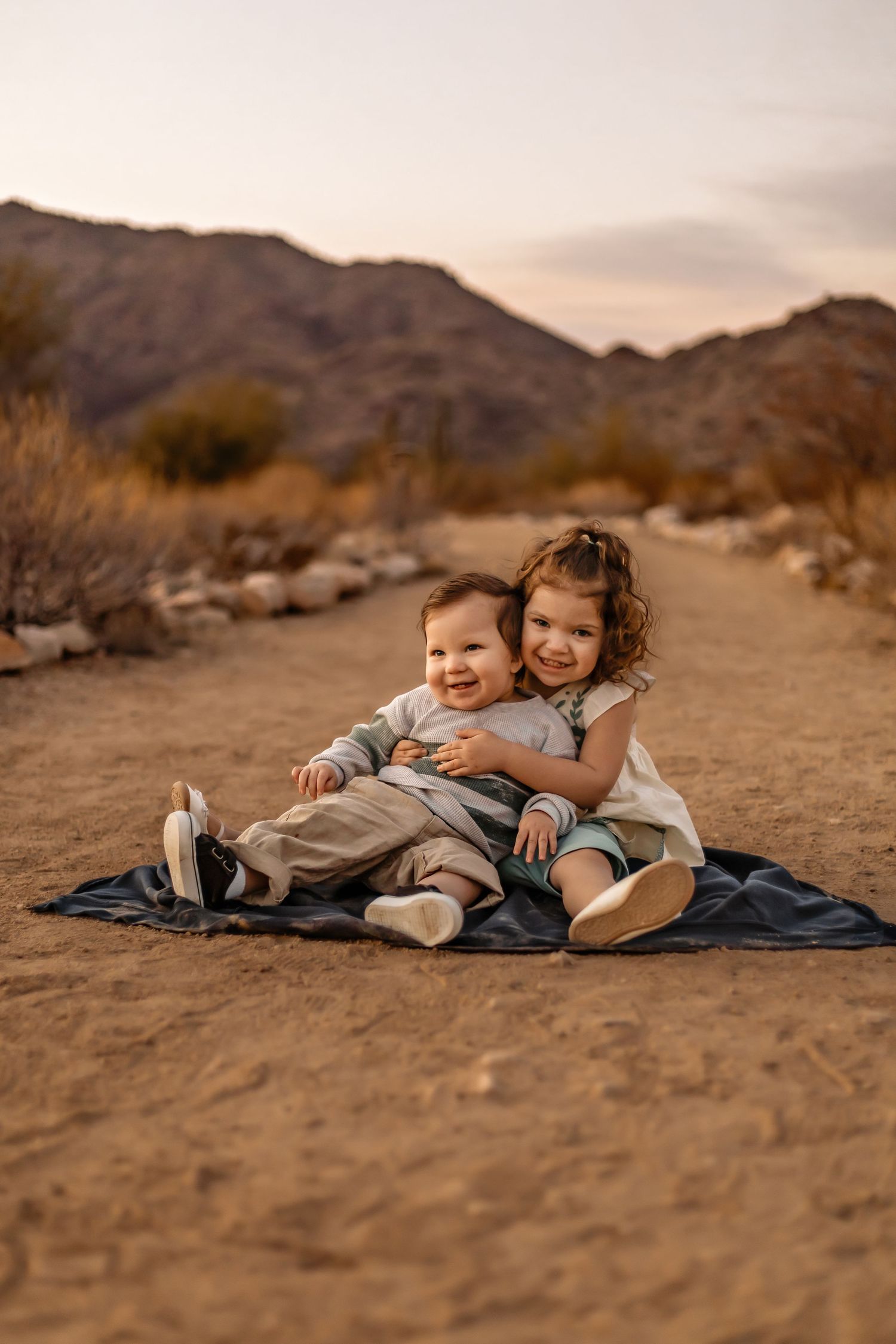 Two children sit together on a blanket in a desert landscape with mountains and warm sunset lighting in the background.