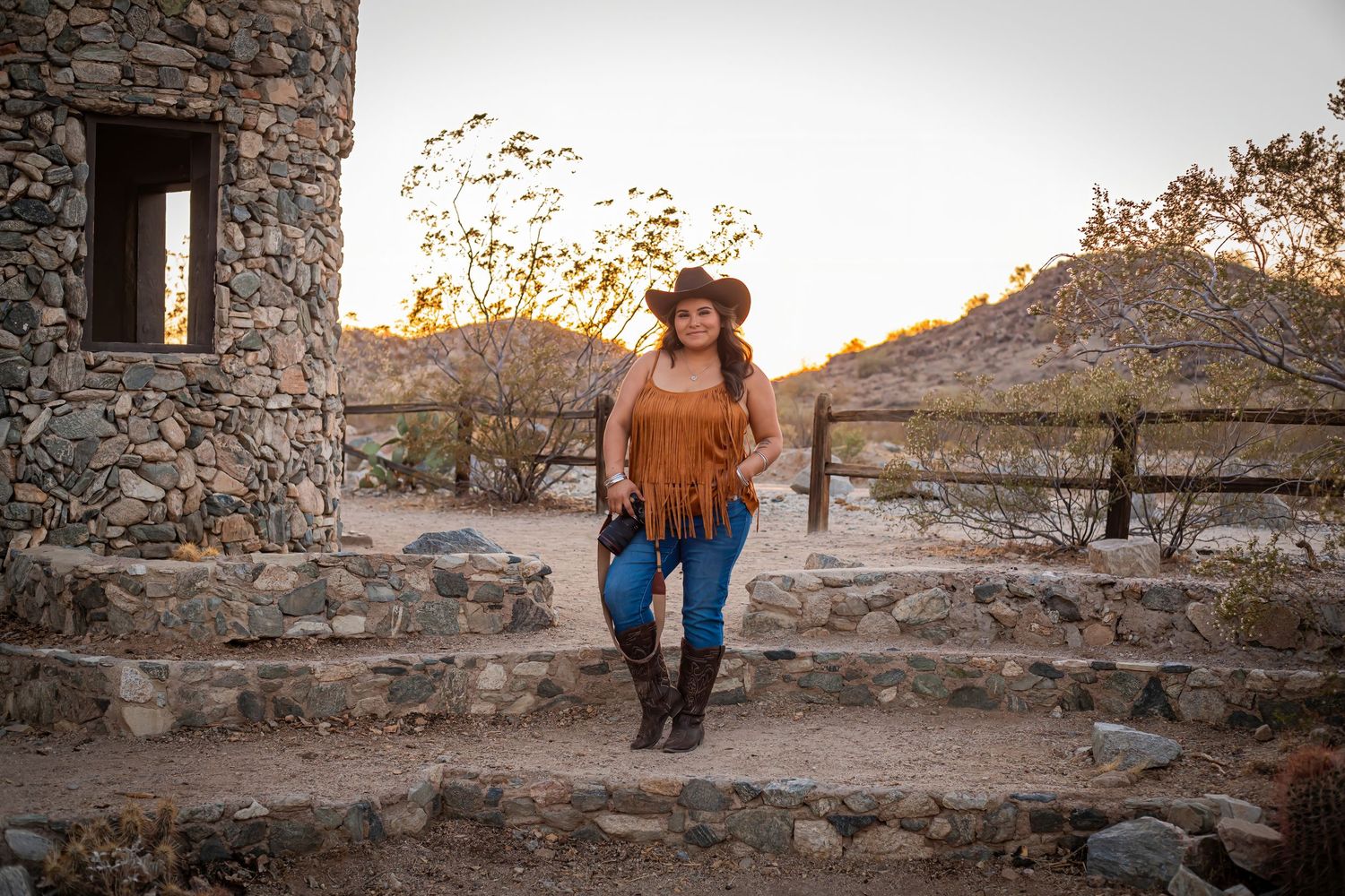 Rustic stone castle wall setting at sunset with someone in Western attire including fringe jacket and cowboy hat.