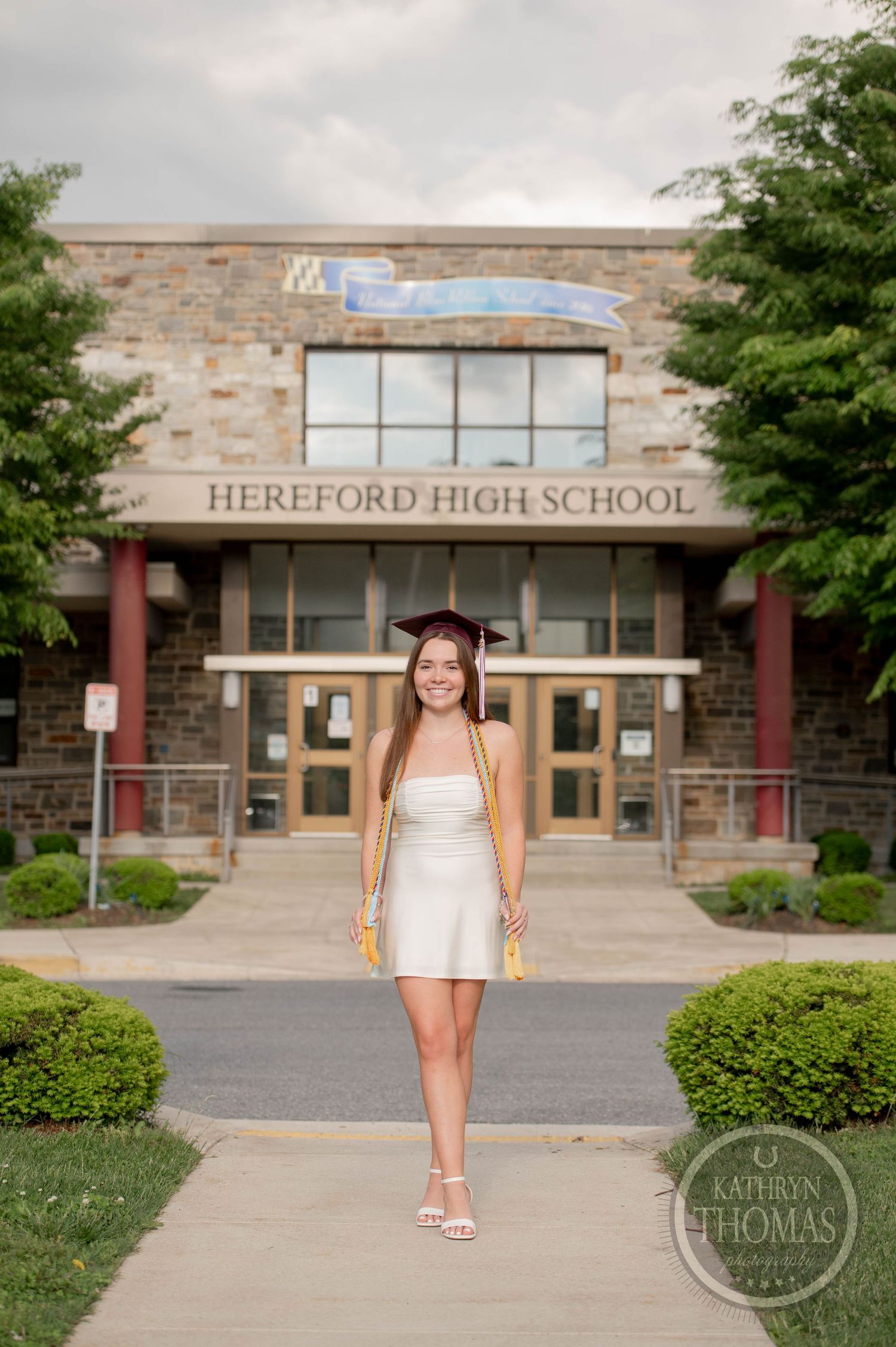 A graduate in a white dress poses in front of Hereford High School's stone facade entrance on graduation day.