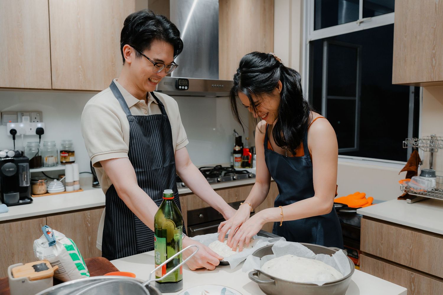 A couple wearing dark aprons preparing food together in a modern kitchen with wooden cabinets.