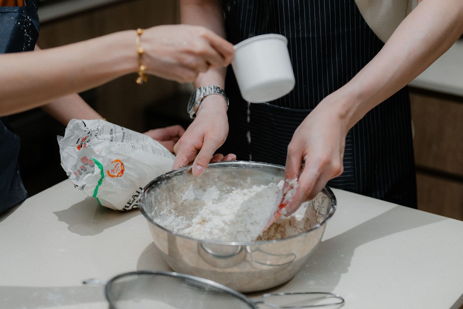 Close up of hands mixing flour and ingredients in a metal bowl on white countertop.