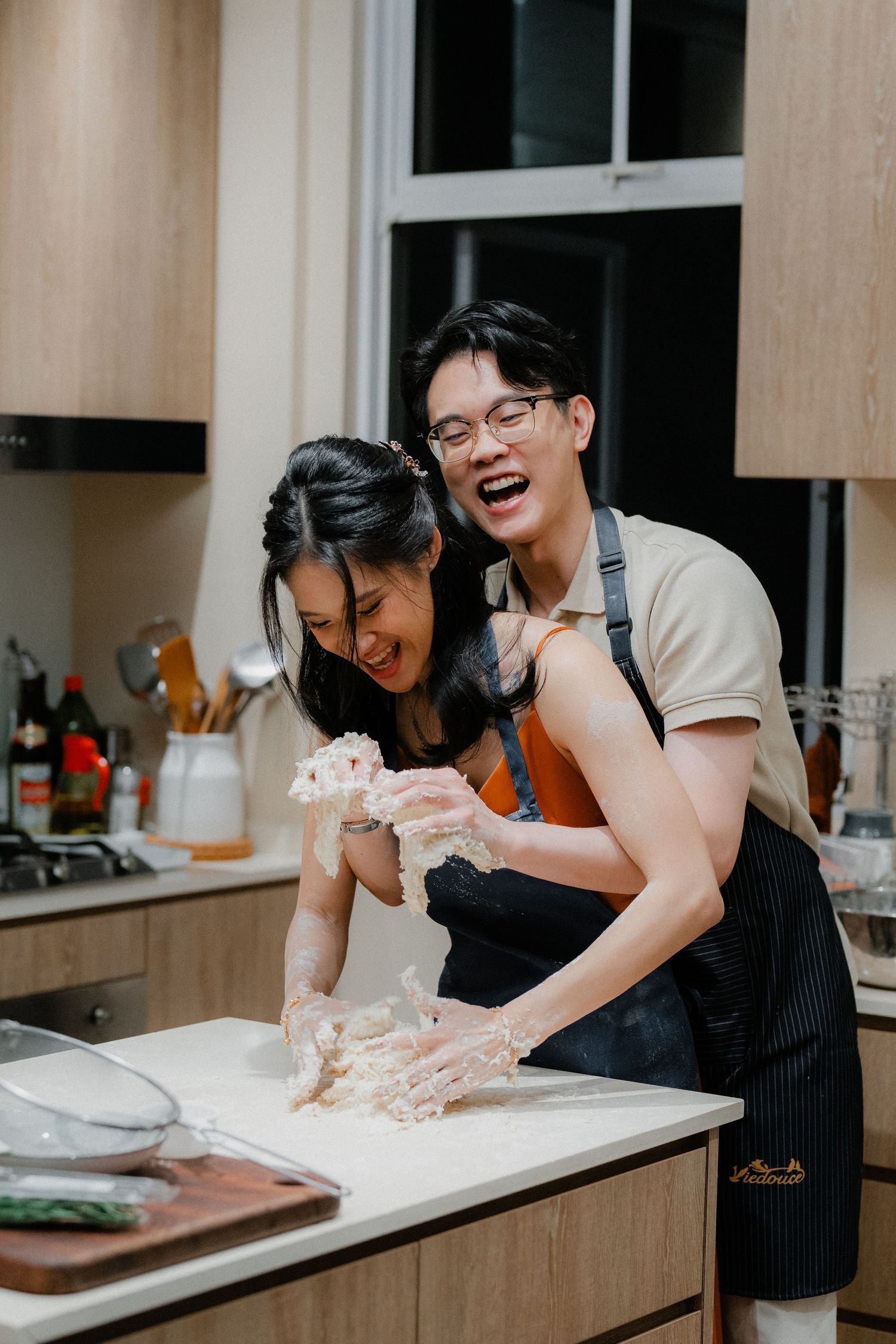 Two people laughing while making dough together in a kitchen with wooden cabinetry.