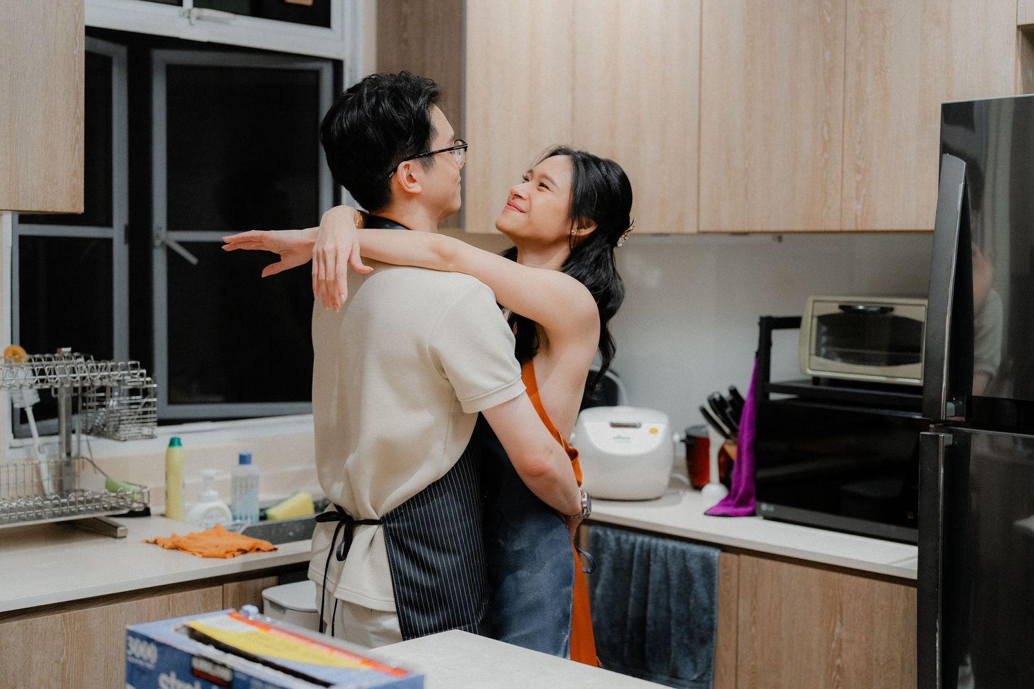 Couple embraces and laughs while baking together in modern kitchen with wooden cabinets.