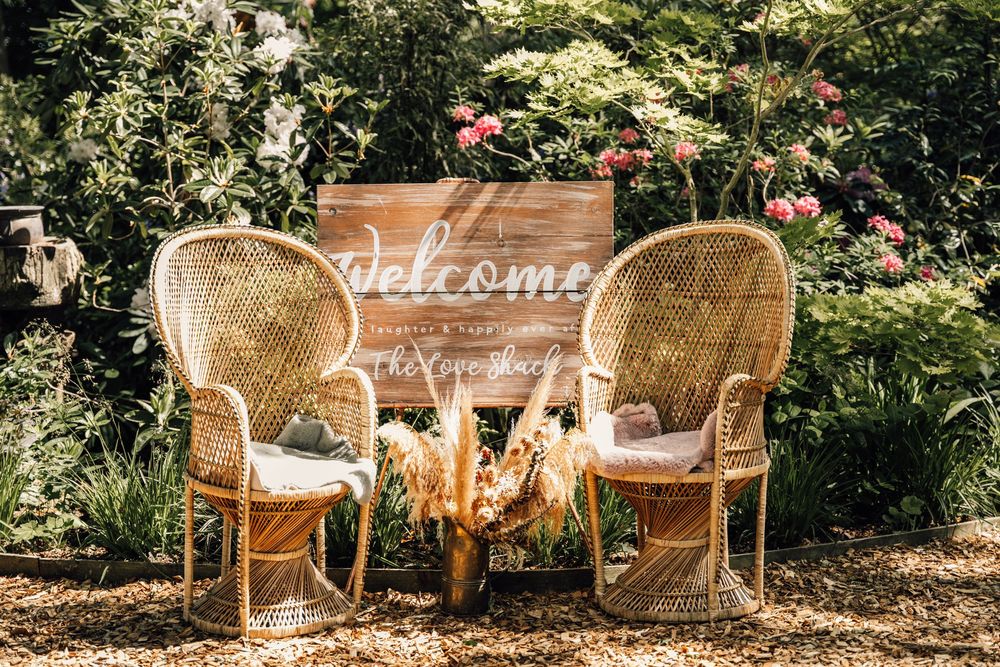 Two vintage wicker chairs and welcome sign arranged in a garden setting with floral decorations.