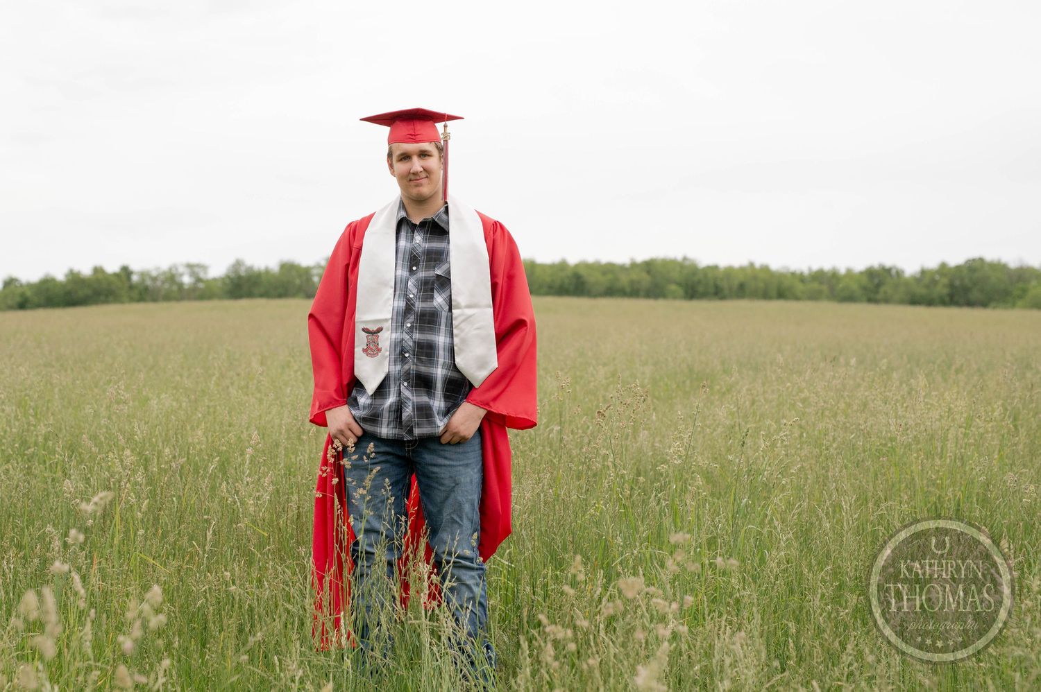 A graduate in red cap and gown stands in a green field on a cloudy spring day.
