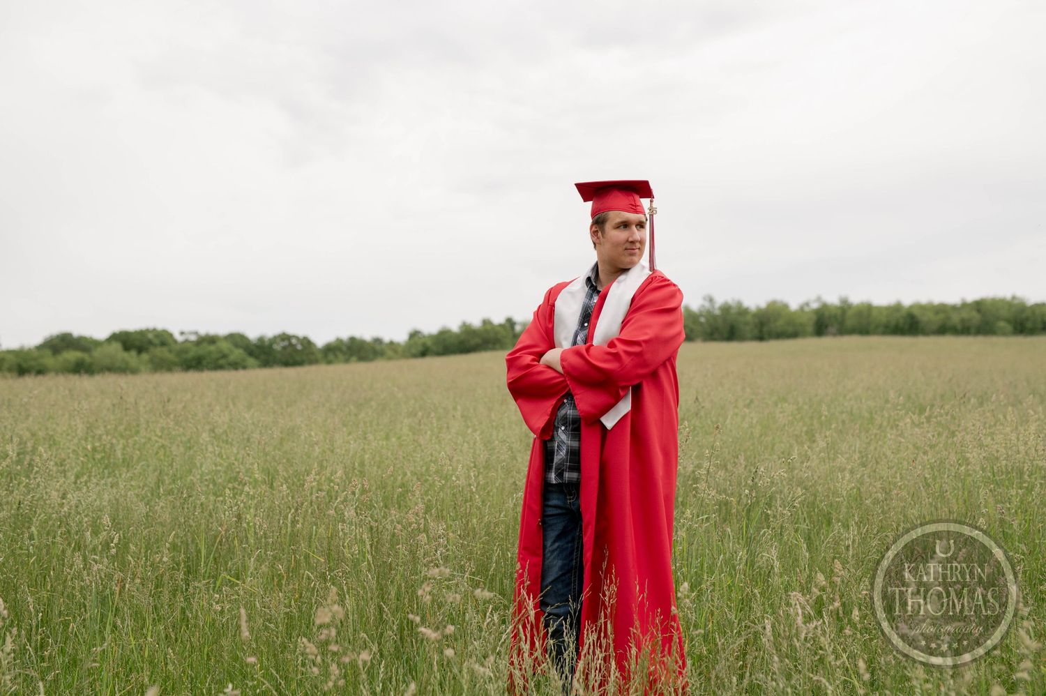 A graduate in a bright red cap and gown stands confidently in a serene meadow of tall grass on an overcast day.