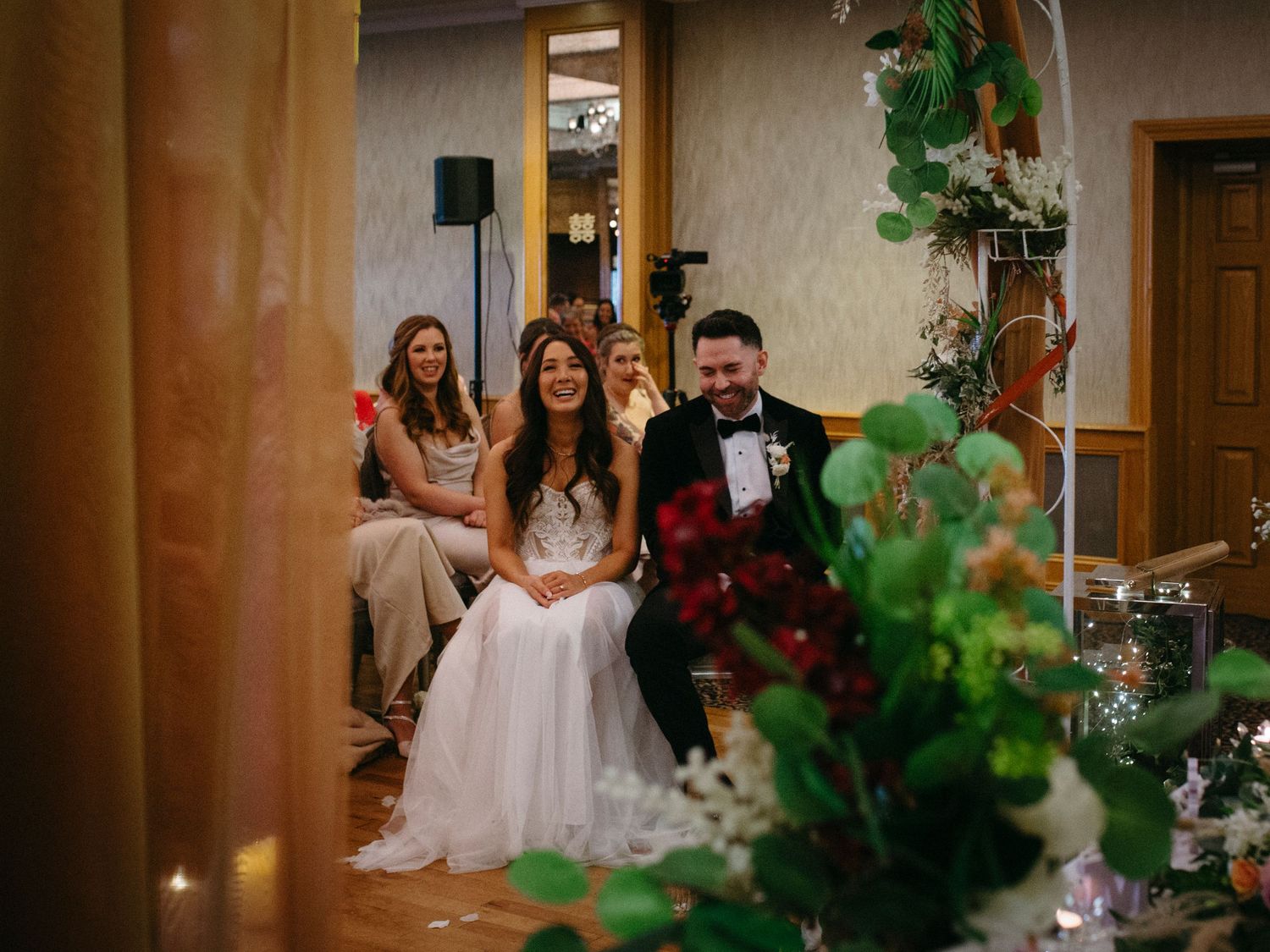 A couple sits together at an indoor venue decorated with green plants during a wedding celebration.