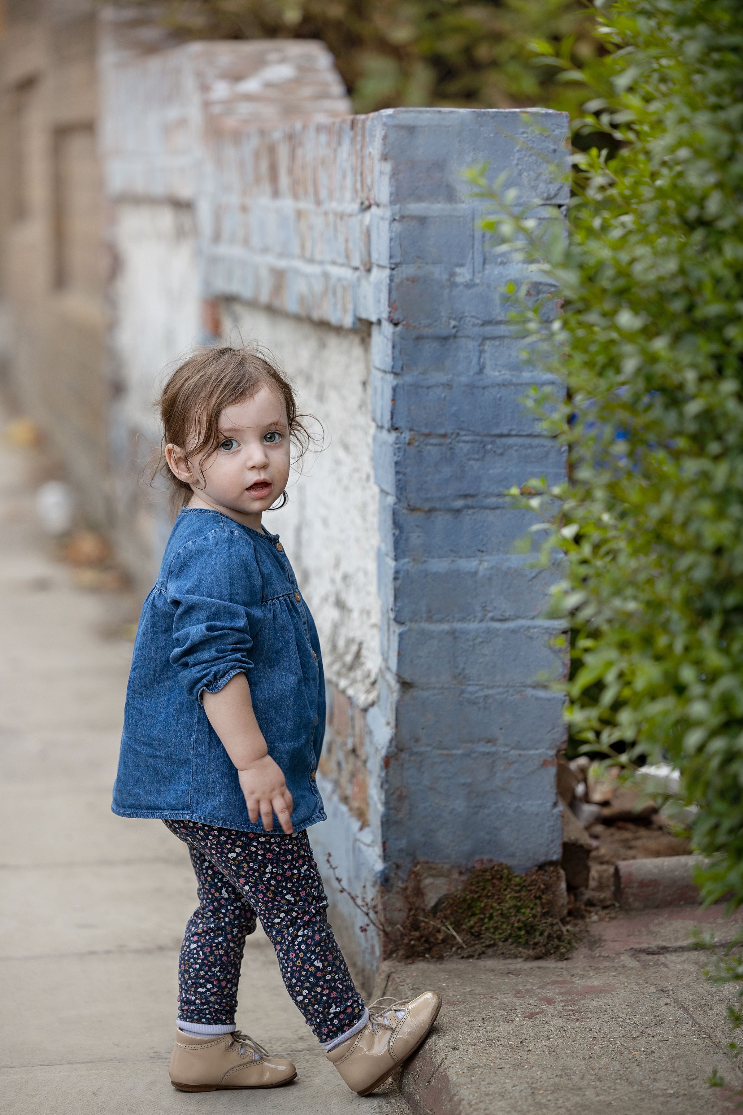Small child in denim jacket and polka dot pants stands on sidewalk near painted brick wall and greenery.