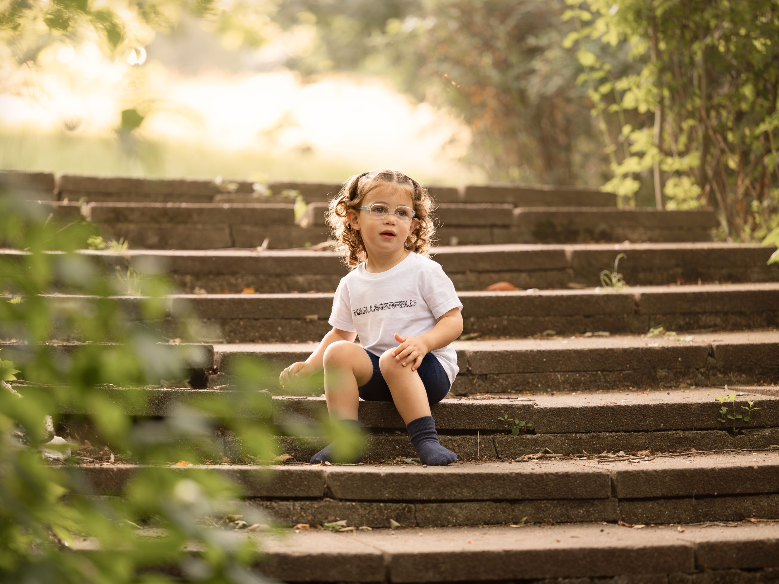 A curly-haired toddler in a white shirt and blue shorts sits on stone steps surrounded by lush greenery in warm sunlight.