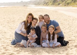 Family portrait of a group sitting together on a sandy beach with coastal cliffs in background.