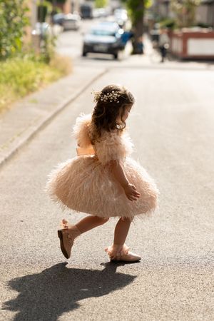 Small child in a fluffy pink tulle dress and floral crown twirls playfully on a sunny residential street.