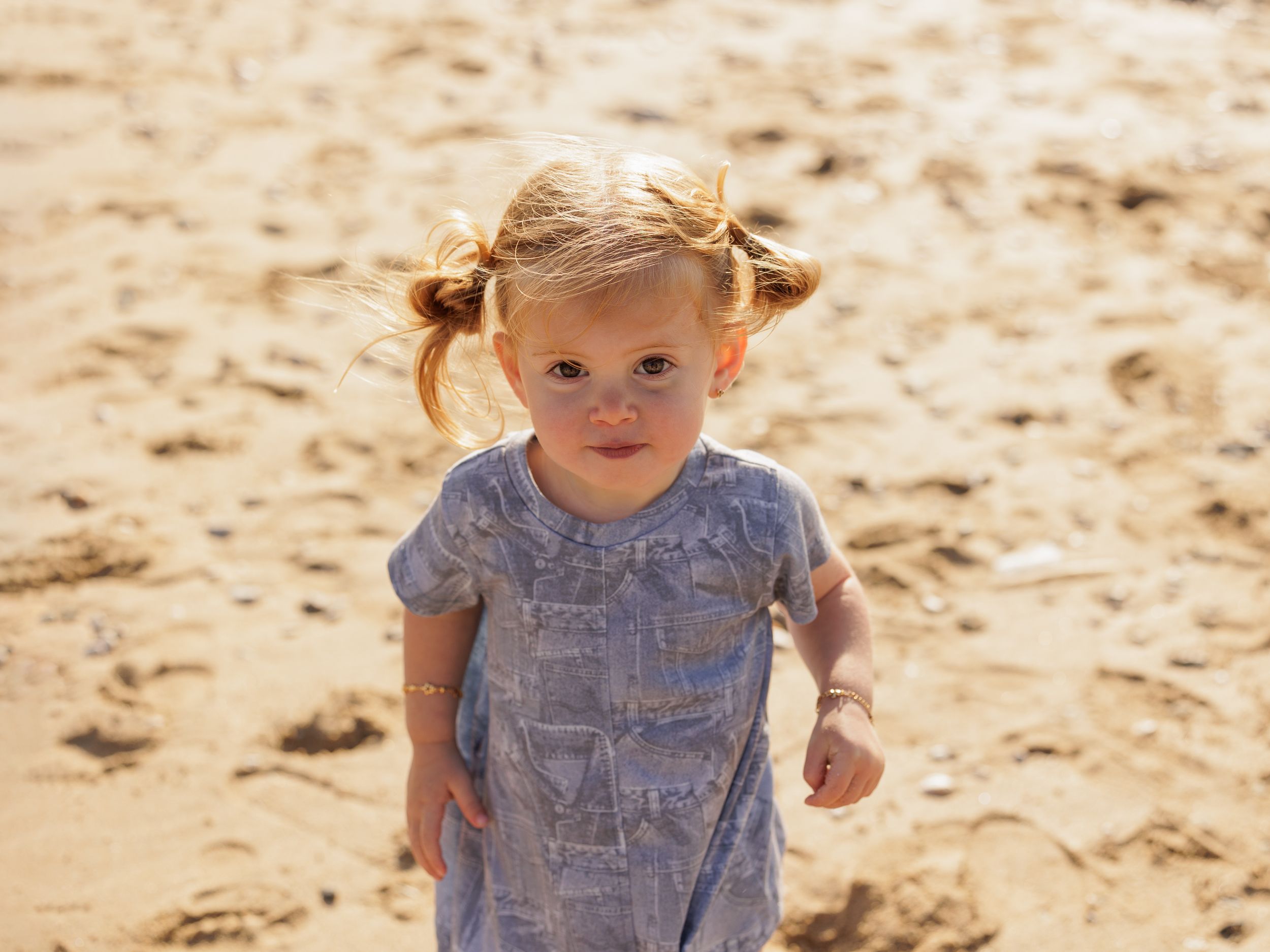 A toddler wearing a light blue dress stands on a sandy beach in warm sunlight.