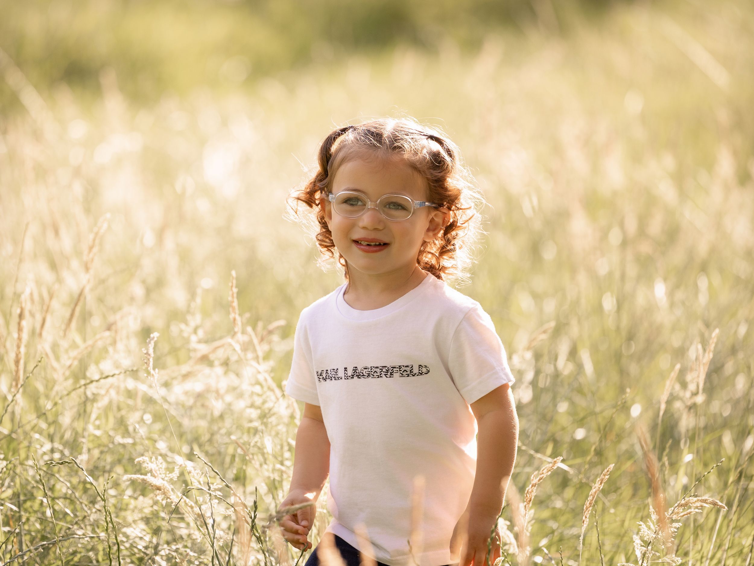 Curly-haired toddler in Karl Lagerfeld shirt smiling in sunny golden meadow grass.