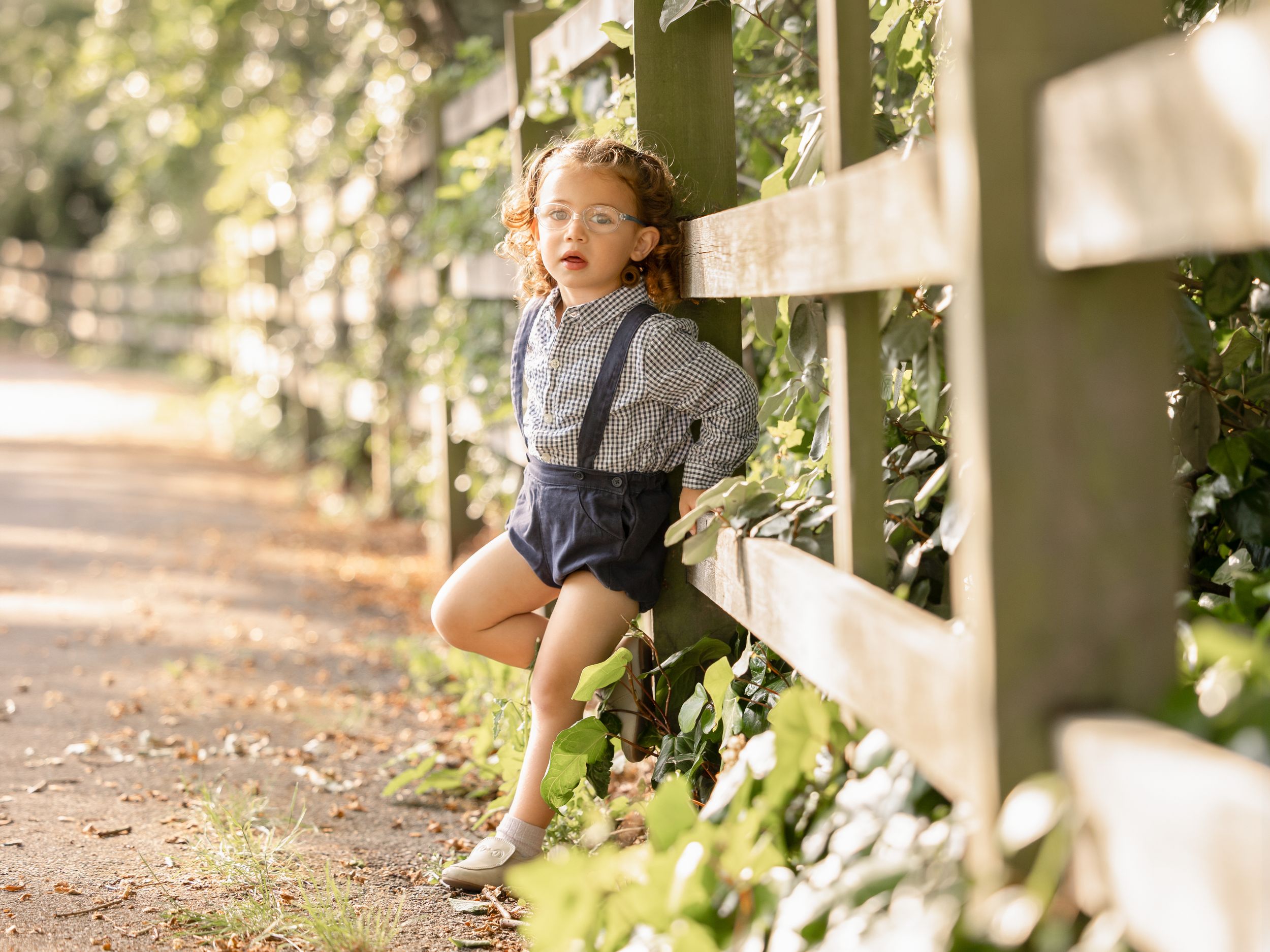 Young toddler in suspenders and shorts leaning against a wooden fence surrounded by ivy on a garden path.