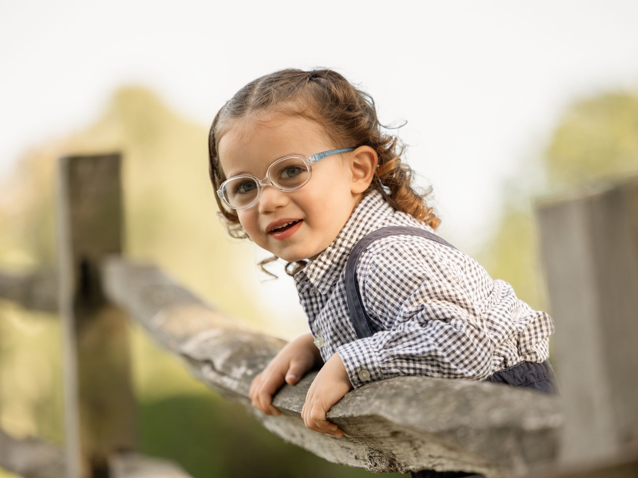 Adorable toddler with curly hair and glasses smiling while leaning on a wooden fence outdoors.