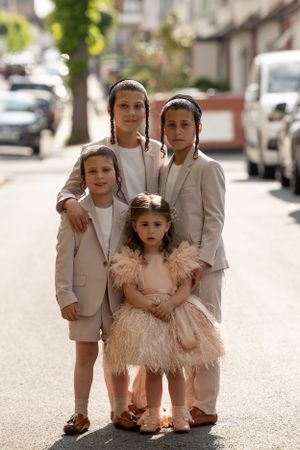 Four figures in matching beige suits pose together on a residential street with one wearing a feathered dress.