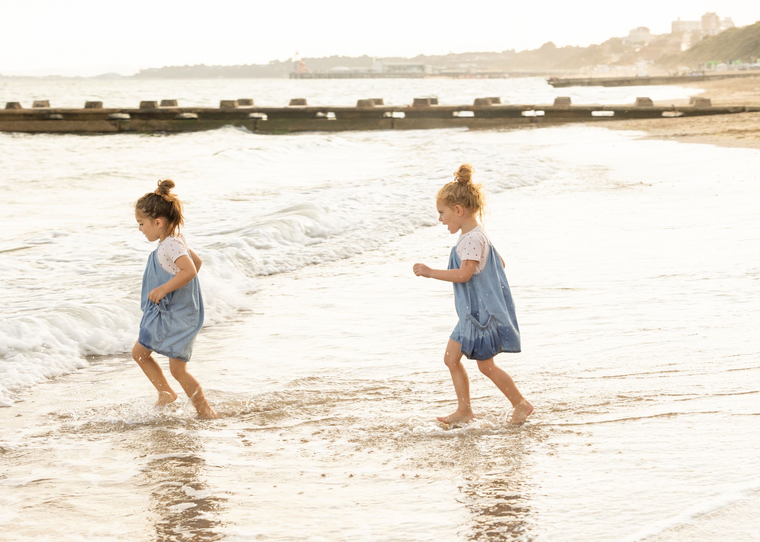 Two children in denim dresses play and splash in shallow ocean waves at sunset along a sandy beach.