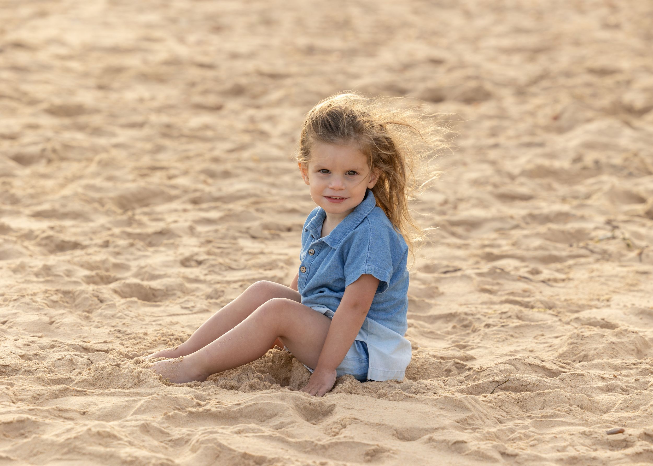 Young child in blue shirt sits on sandy beach during golden hour with wind-blown hair.