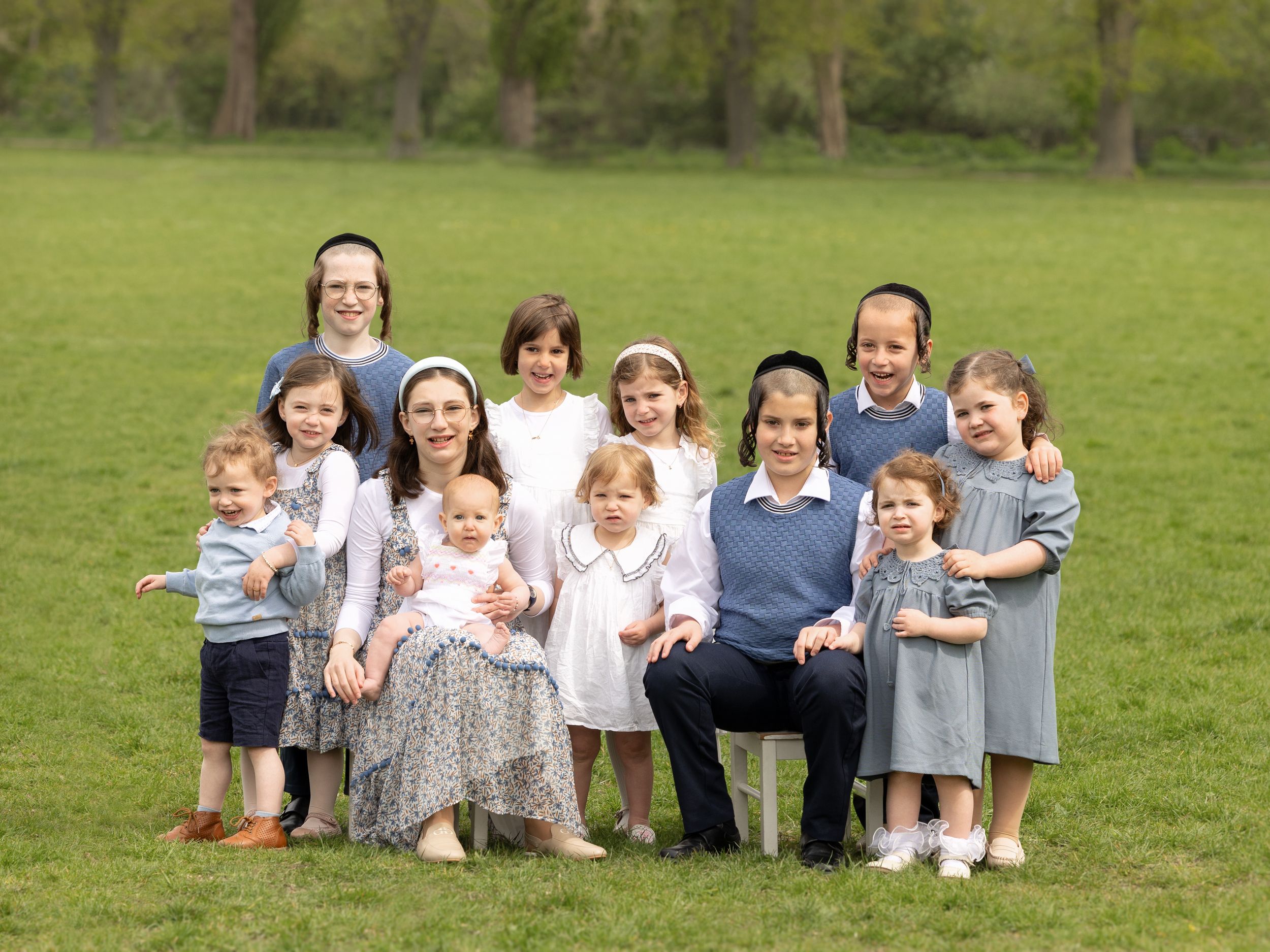 A group of young children and adults pose together on a grassy lawn with trees in the background.