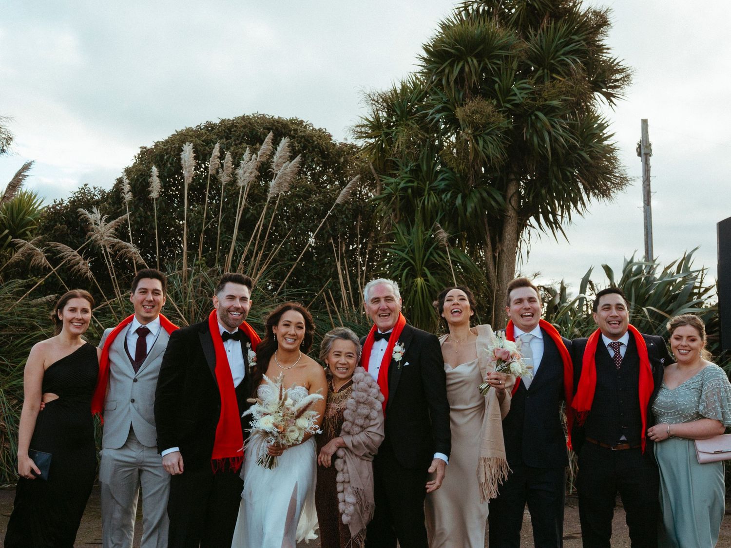 A wedding party poses together outdoors in front of tropical foliage and pampas grass.