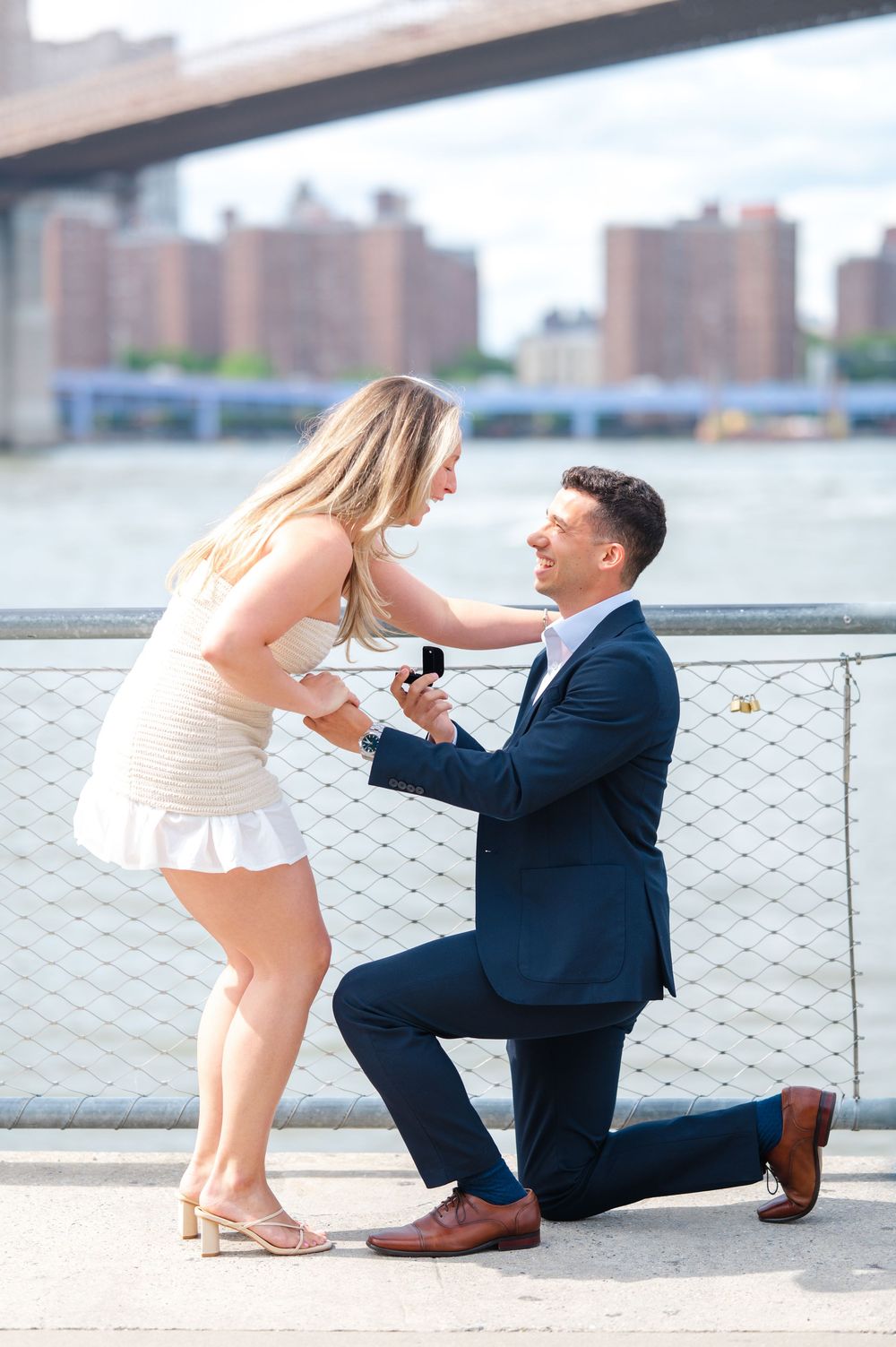 A romantic proposal with a man in a suit kneeling while holding a ring in front of a cityscape at Brooklyn Bridge Park while the woman cries