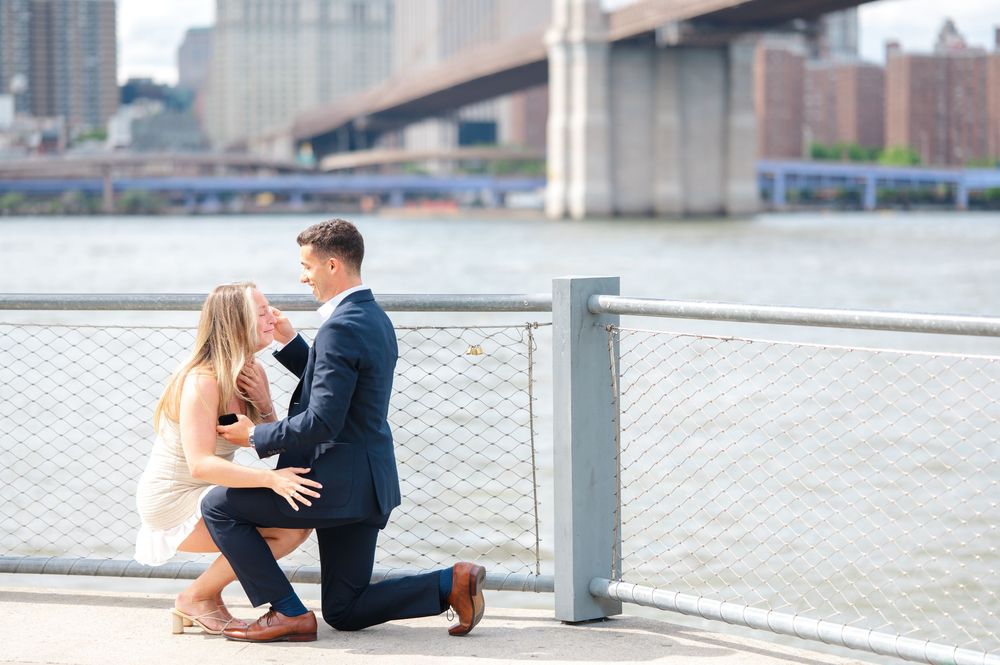 Two people share a tender moment during their proposal at the Brooklyn Bridge Park with a cityscape and bridge visible in the background.