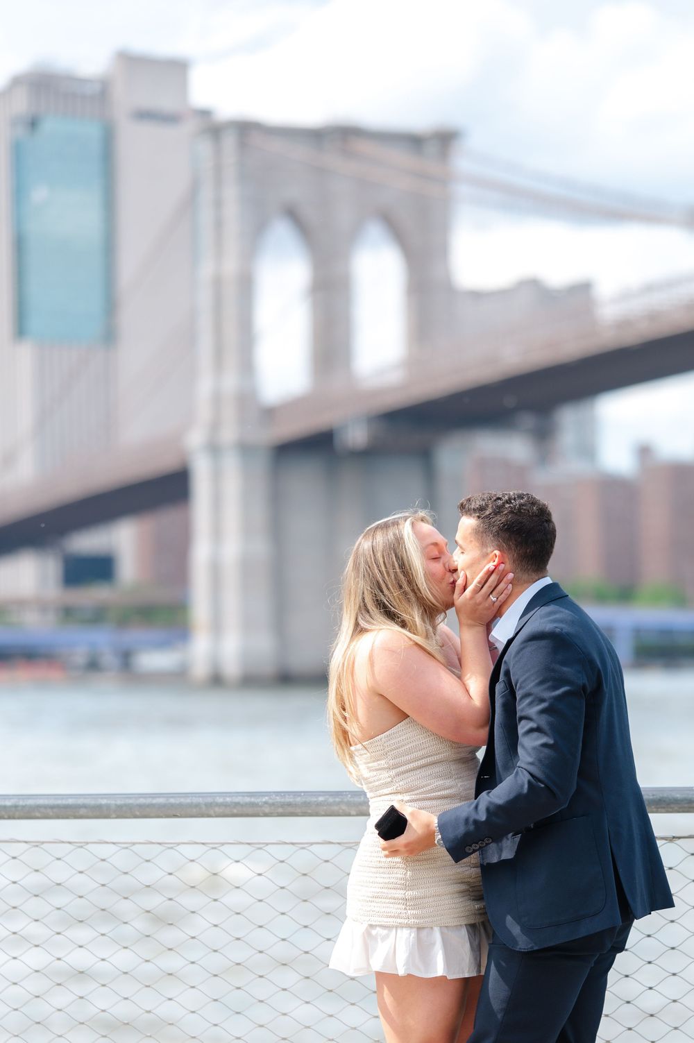 Couple sharing a romantic kiss in front of the Brooklyn Bridge on a sunny day after their NYC proposal.