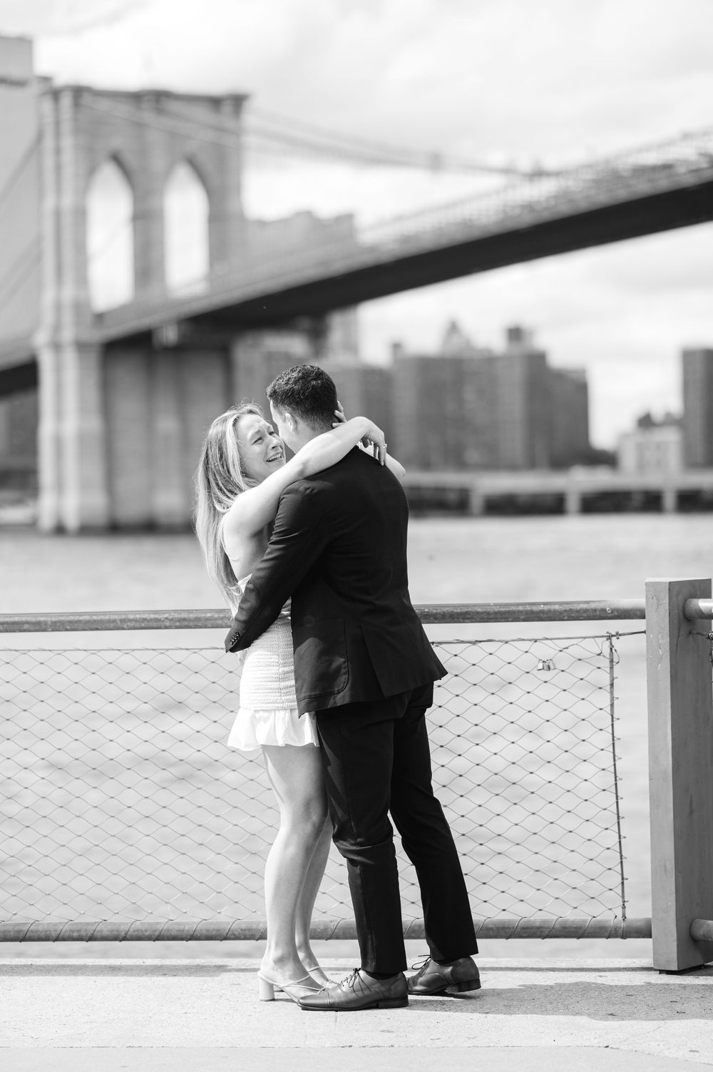 Romantic black and white photo of a couple embracing at the Brooklyn Bridge Park with city skyline in background. Photo by Joliette Marie.