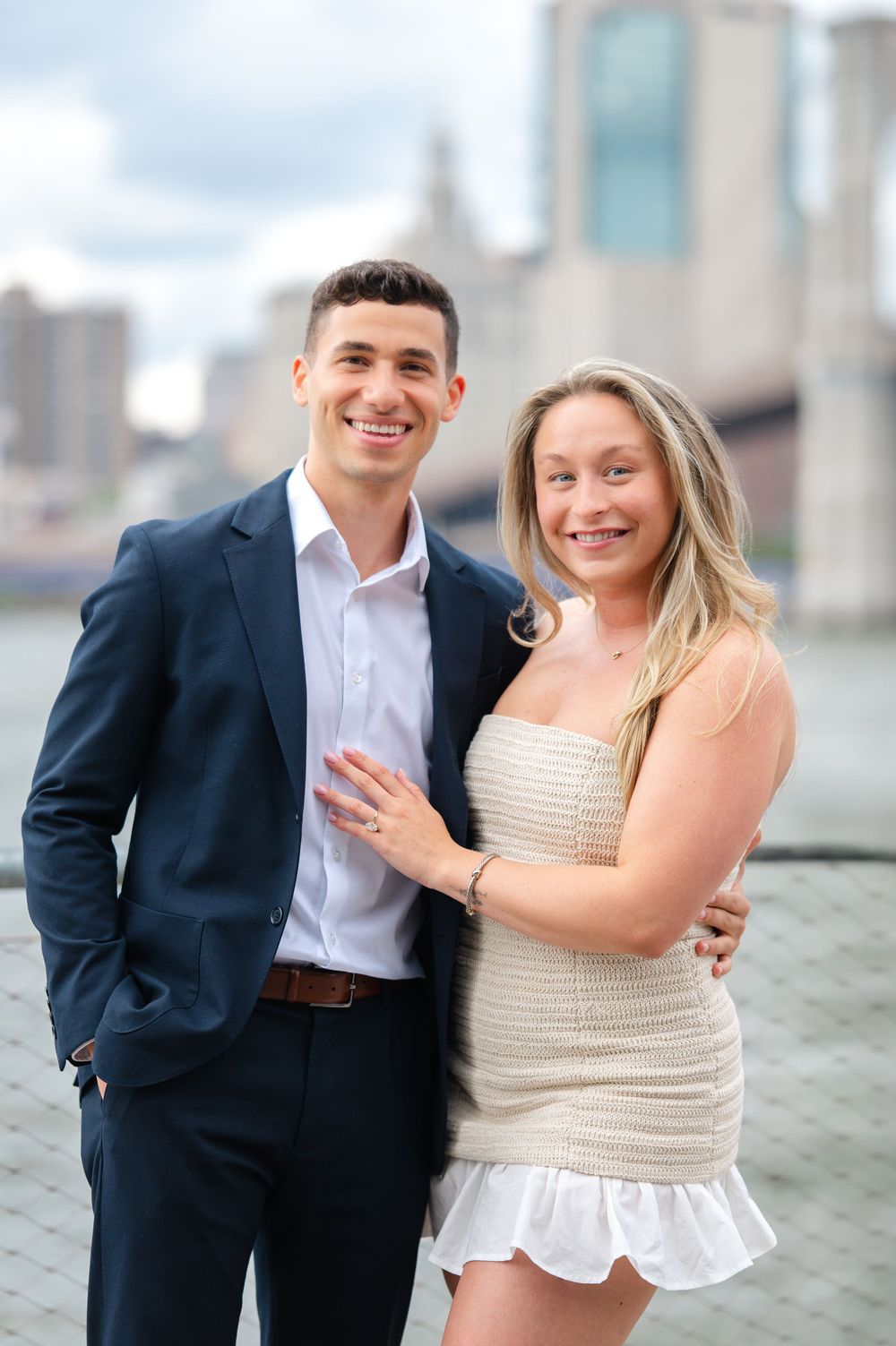A happy couple poses for engagement photos by the Brooklyn Bridge waterfront after their NYC surprise proposal.