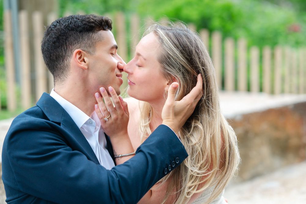 A romantic couple in formal attire share a tender embrace outdoors with a wooden fence in the background.