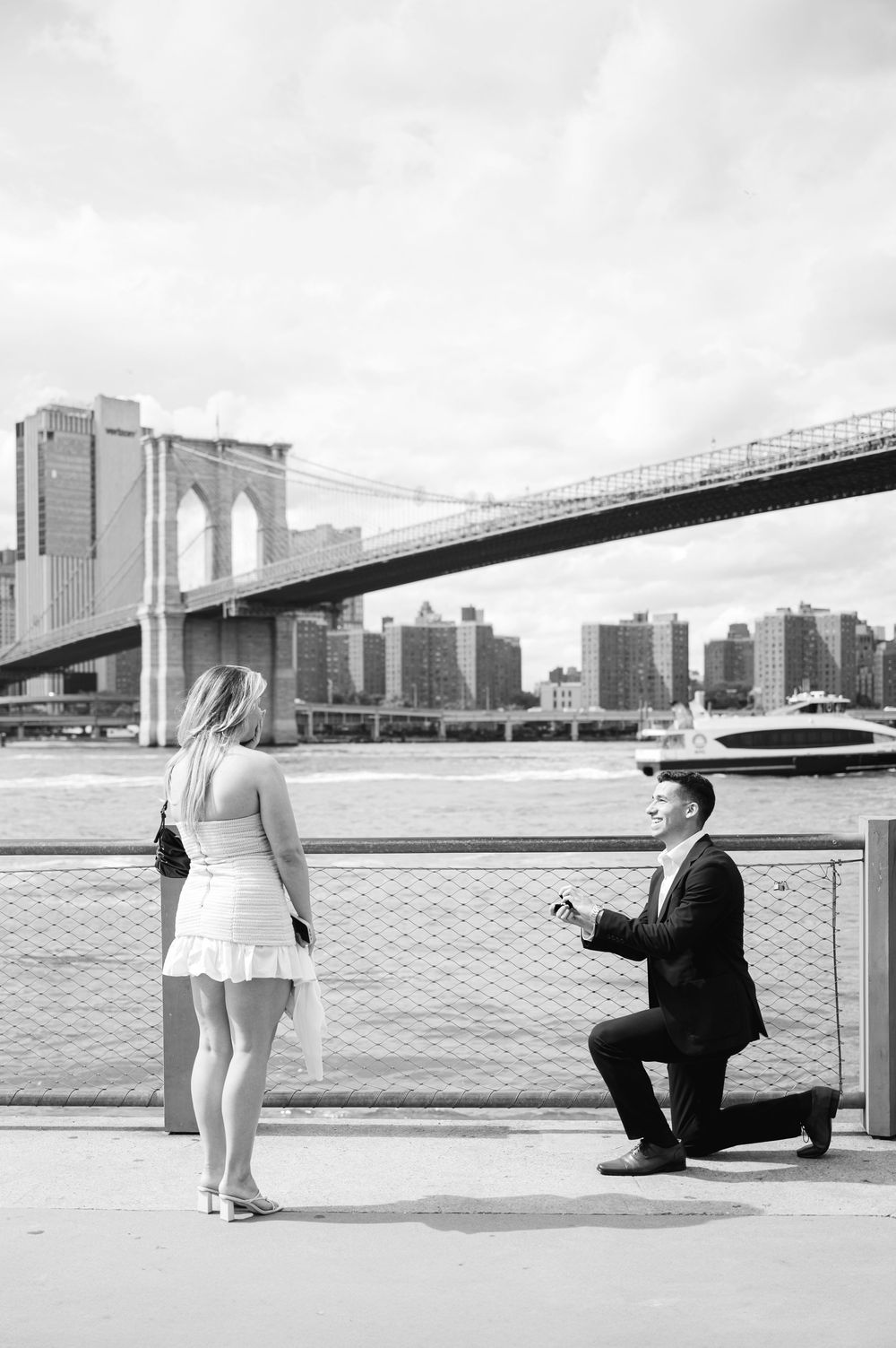 A black and white photo shows a romantic marriage proposal scene with the Brooklyn Bridge in the background.