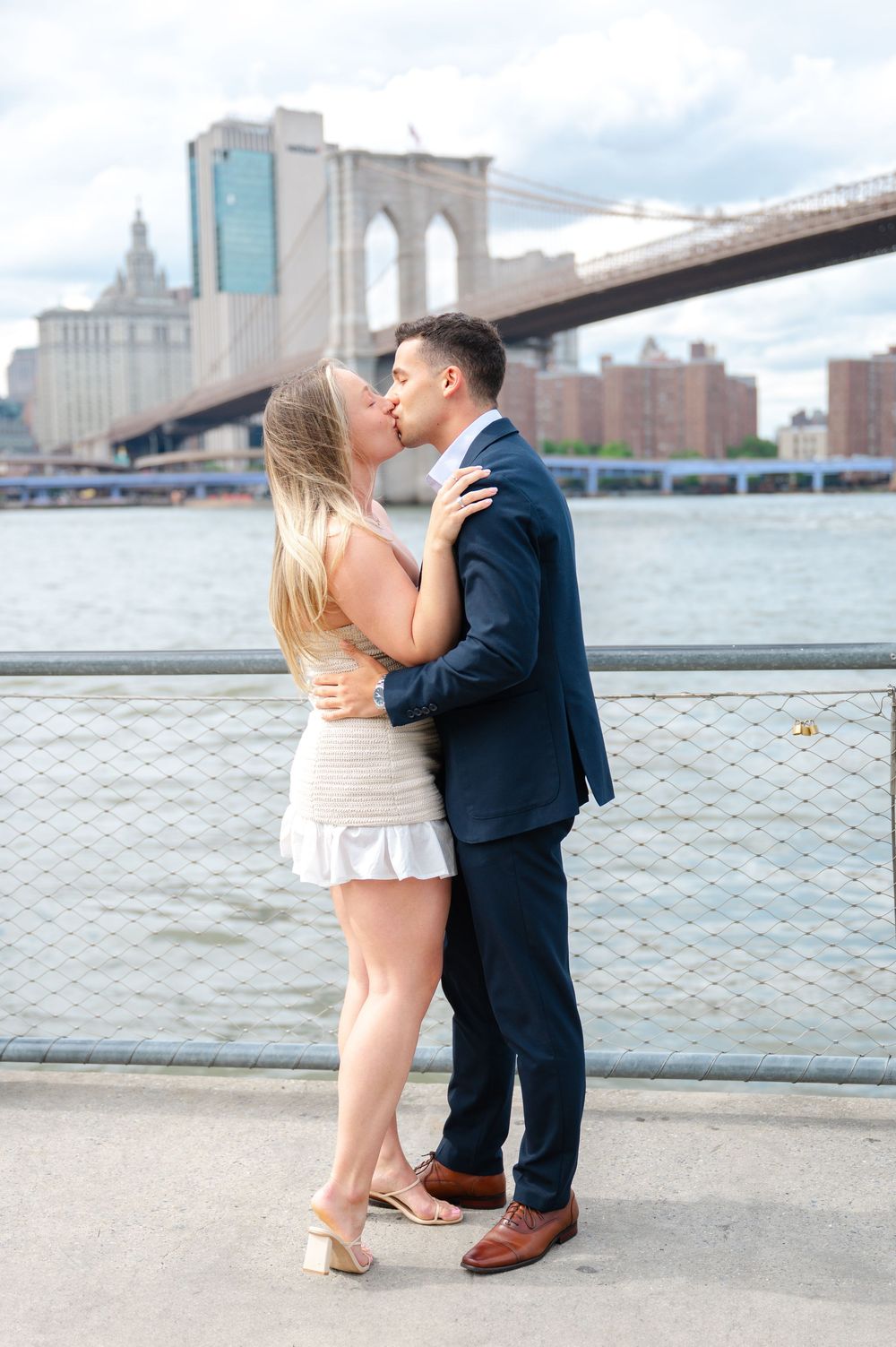 Romantic couple embracing near Brooklyn Bridge with Manhattan skyline and East River in background.