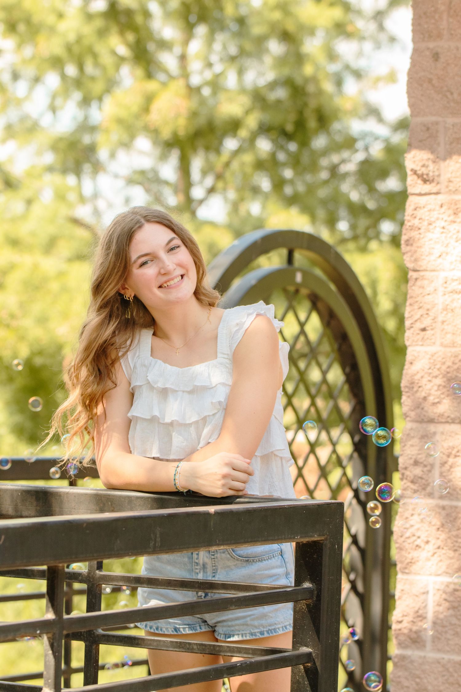 Young woman in white ruffled tank top and denim smiles while leaning on a decorative garden arbor on sunny day.
