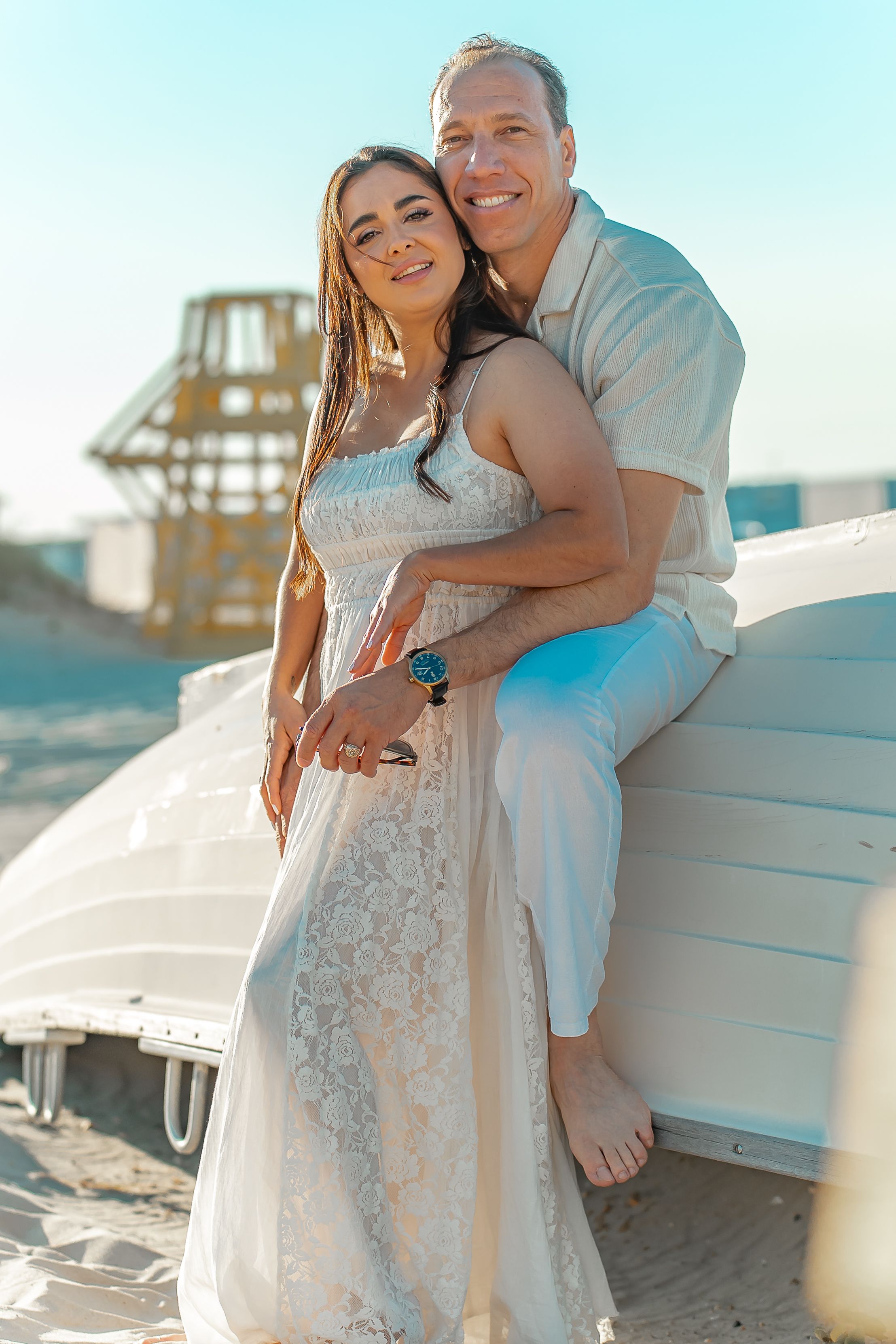 A couple in white clothing embracing by beach lounge chairs during a sunny day.