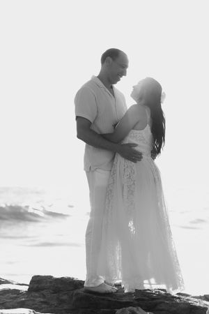 A black and white portrait of a couple in white attire embracing on coastal rocks.