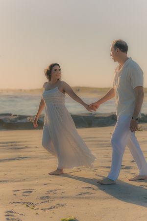 A couple in white clothing dances romantically on a beach at sunset.