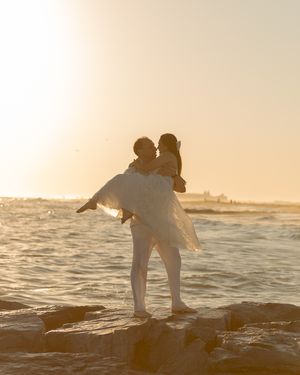 A silhouetted couple in white clothing dancing on rocks by the ocean at sunset.