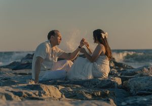 A couple in white outfits posing romantically on coastal rocks with ocean waves.