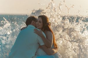 A couple sharing a romantic moment with ocean waves splashing behind them at sunset.