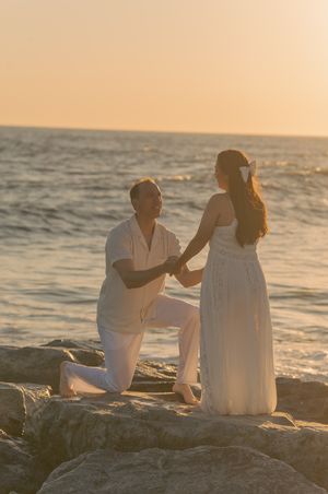 A couple in white clothing sitting on coastal rocks during a golden sunset.