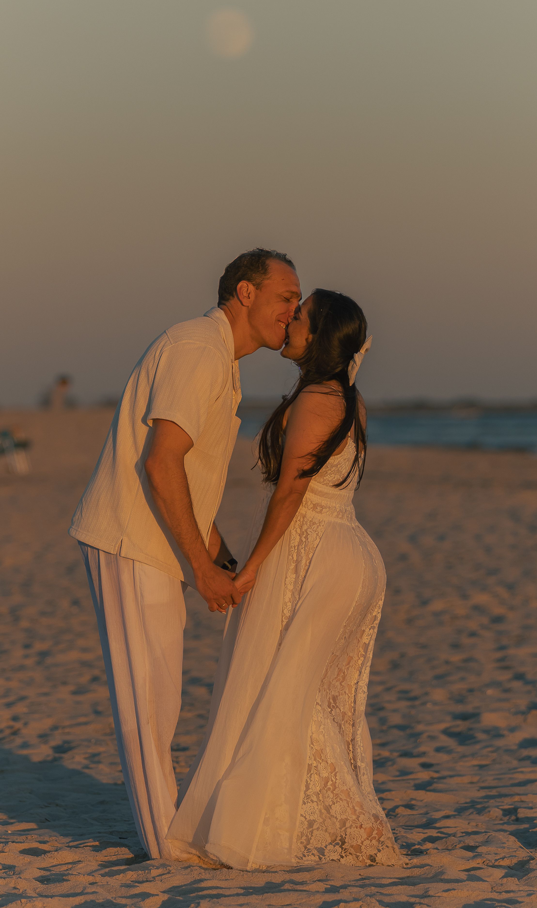 A romantic couple in white outfits posing together on sandy beach at dusk.