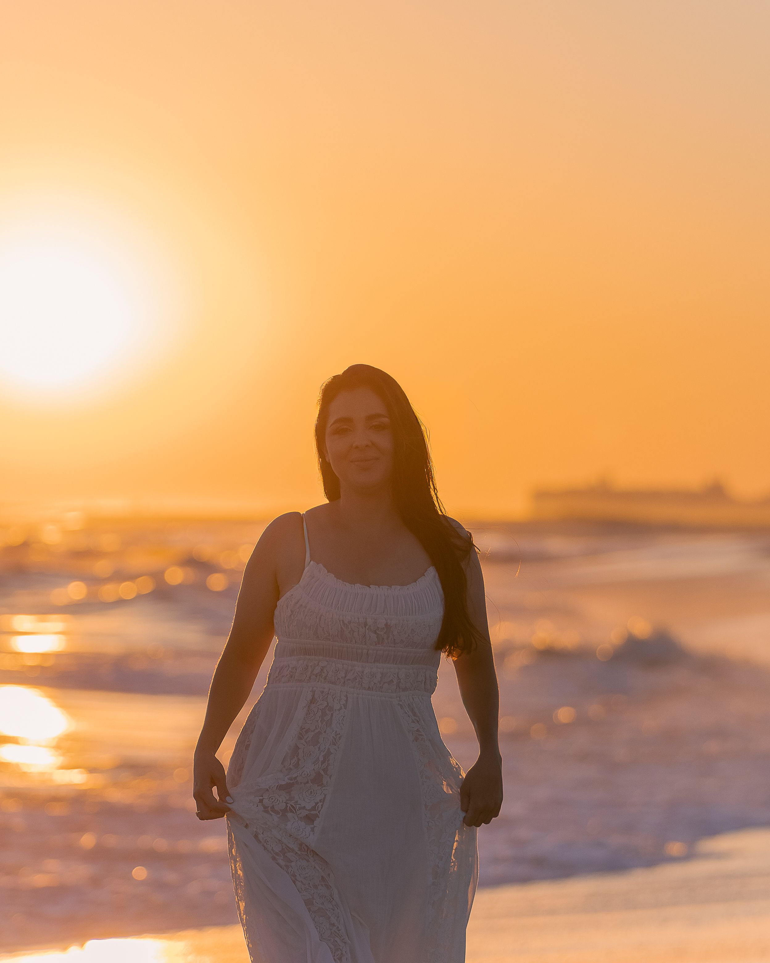 A romantic beach photoshoot at sunset with a couple in white attire against an orange sky.